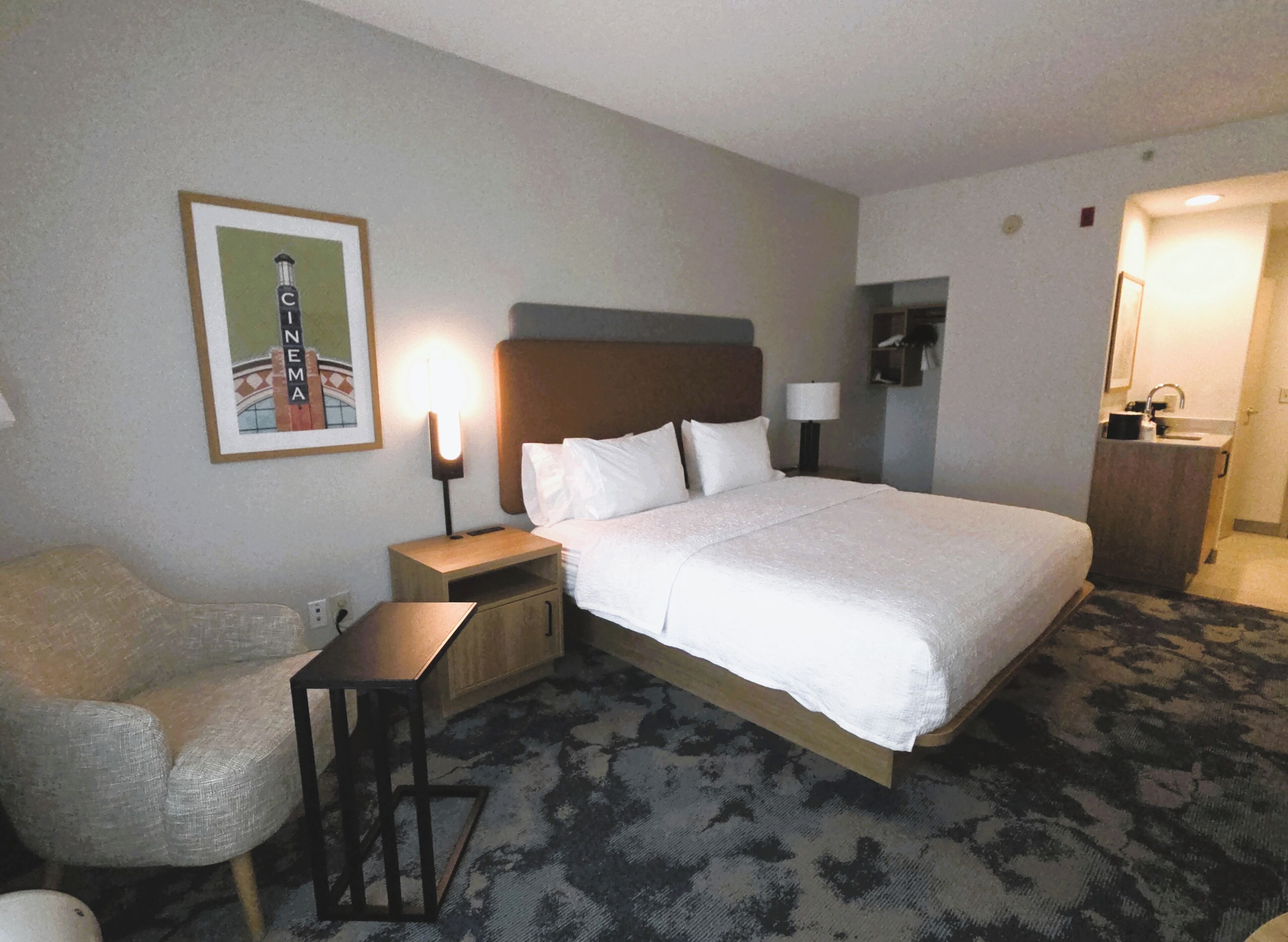 Habitación de hotel con cama, una silla, mesita de noche, lámpara, un cuadro y baño.
