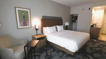Habitación de hotel con cama, una silla, mesita de noche, lámpara, un cuadro y baño.