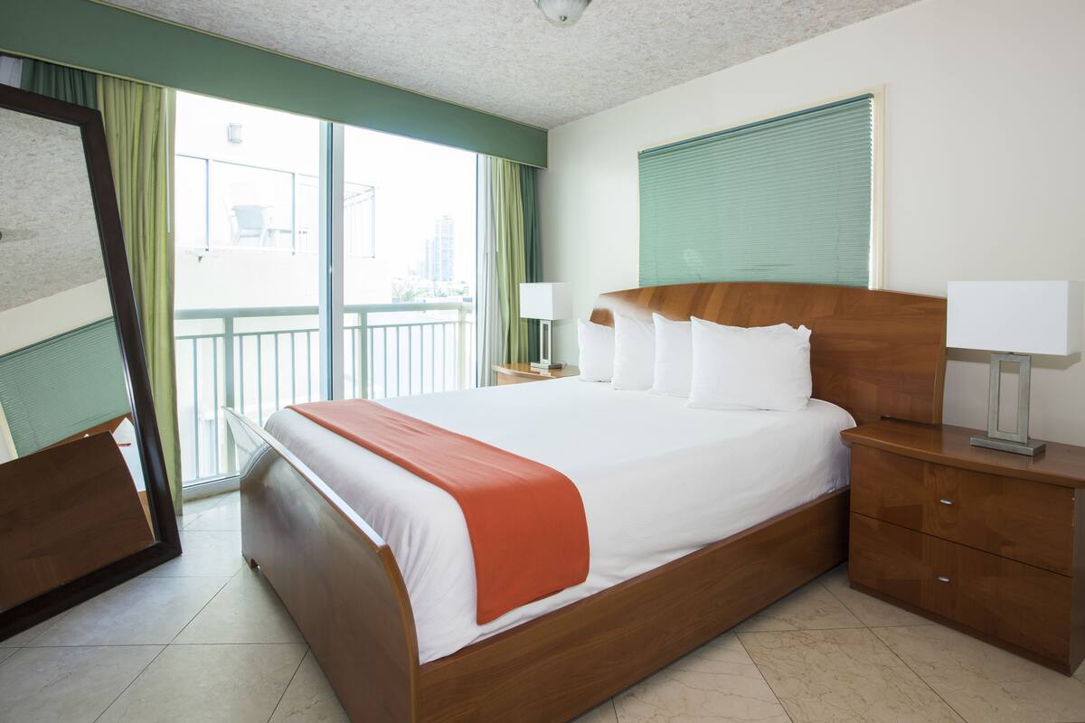 Habitación de hotel con una cama grande, una mesita de noche con lámparas, un espejo y vistas al balcón exterior.