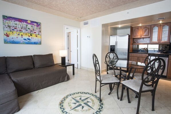 A living room with a sofa, a dining table with chairs, a kitchen area with cabinets, and a wall with a colorful painting.