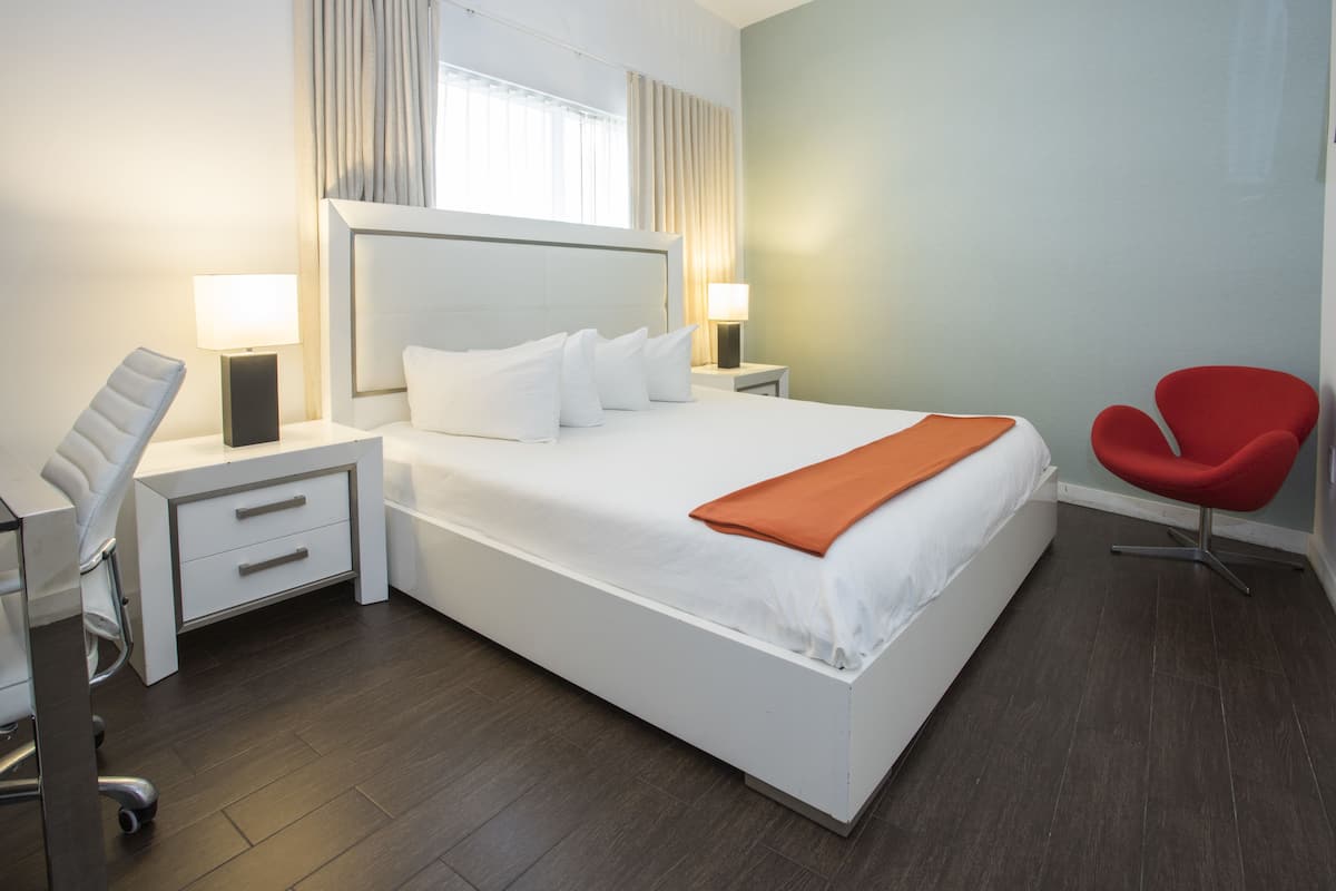 Una habitación de hotel moderna con una cama grande, dos mesitas de noche con lámparas, una silla roja y un escritorio con silla.