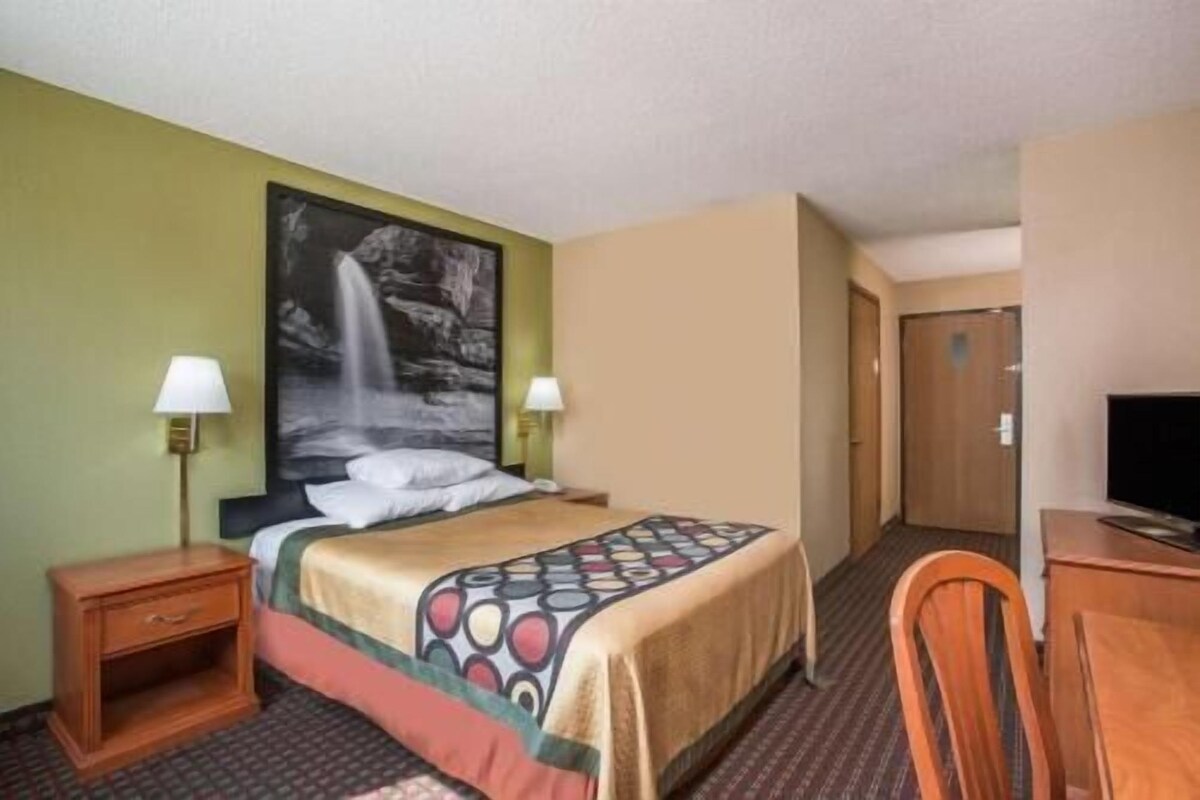 Habitación de hotel con una cama, dos lámparas de noche, un cuadro de una cascada, una mesita de noche de madera, un escritorio con una silla y un televisor.