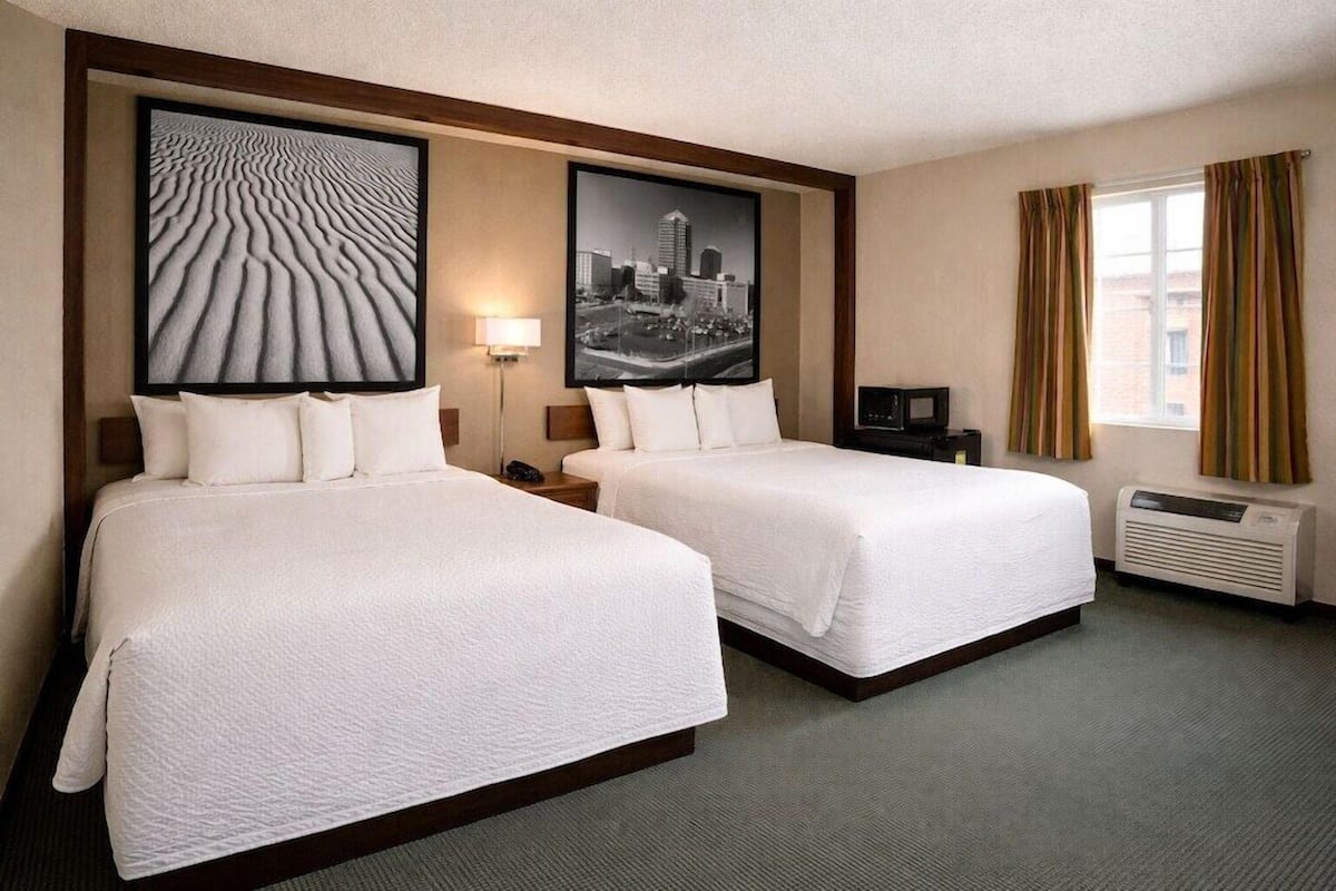 A hotel room with two beds, a cityscape artwork, a lamp, an air conditioning unit, and a television.