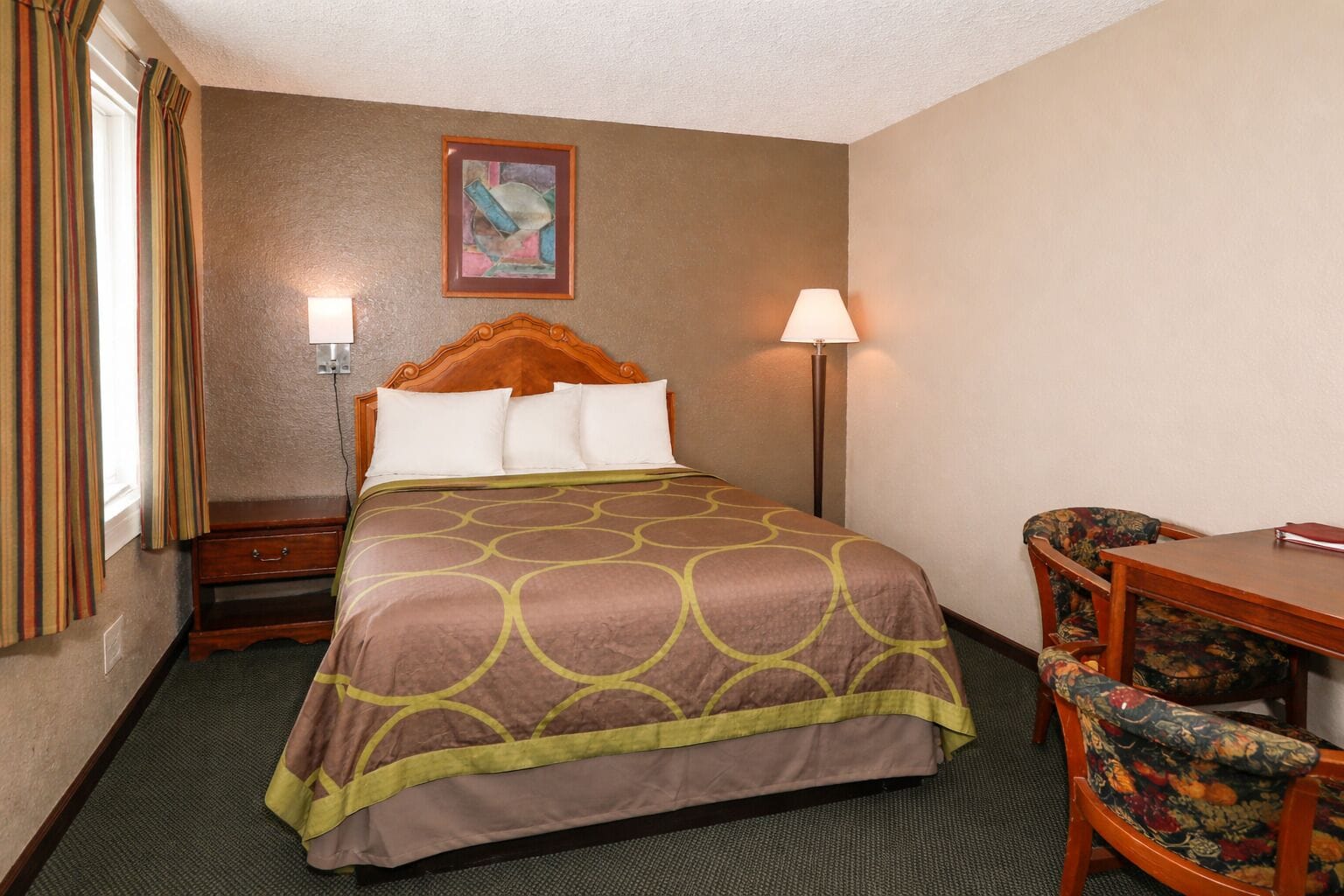 A hotel room with a bed, a desk, a chair, a lamp, a painting on the wall, and a window with curtains.