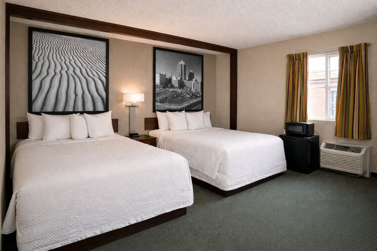 A hotel room with two beds, a nightstand, a lamp, a television, and an air conditioning unit.