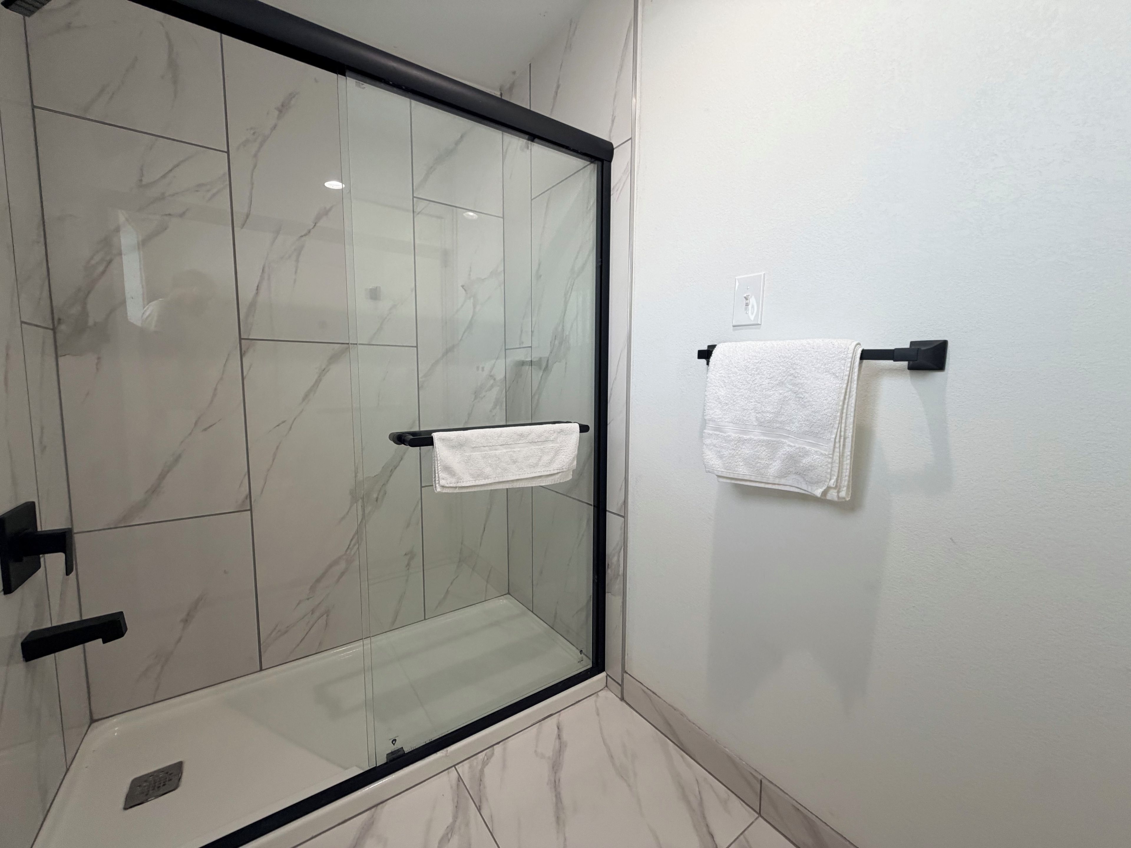 Un baño con cabina de ducha de vidrio, suelo de azulejos blancos y un perchero con dos toallas blancas.