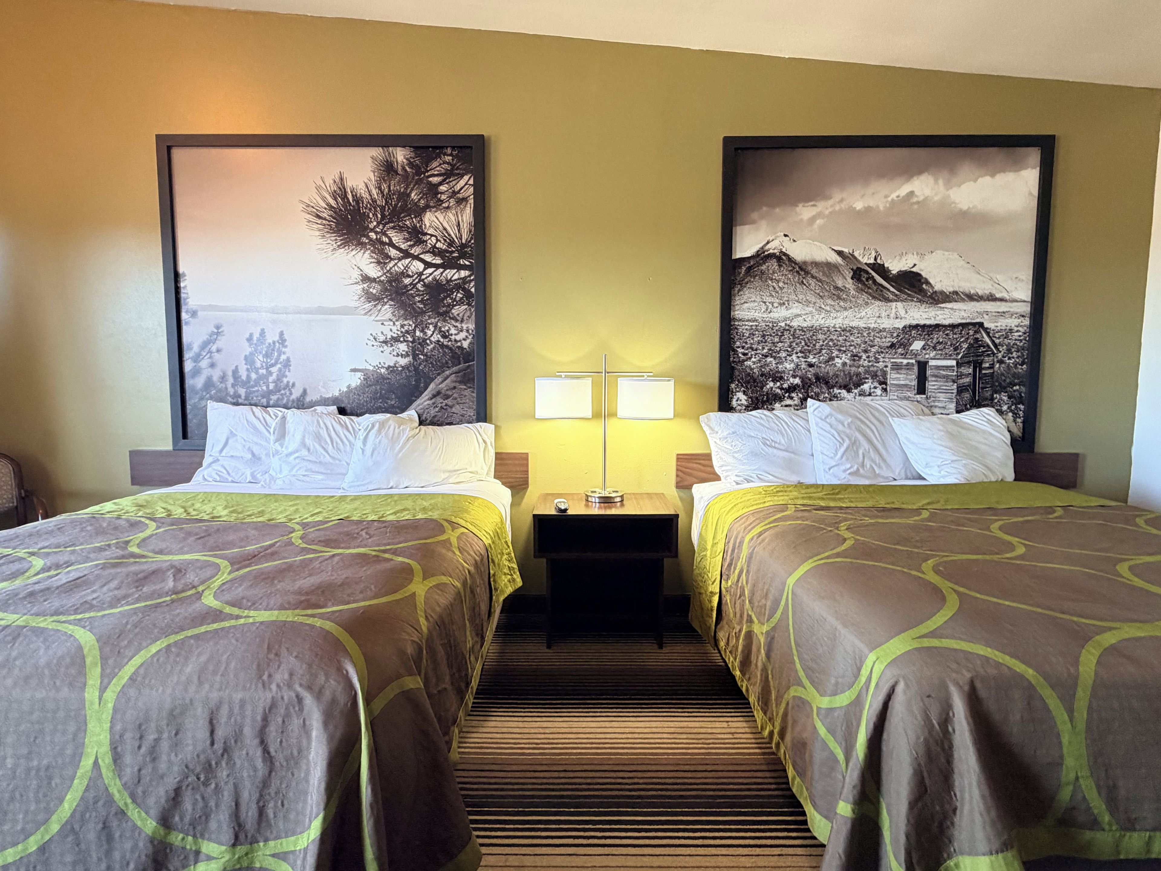 A hotel room with two beds, a nightstand with lamps, and large landscape paintings on the walls.