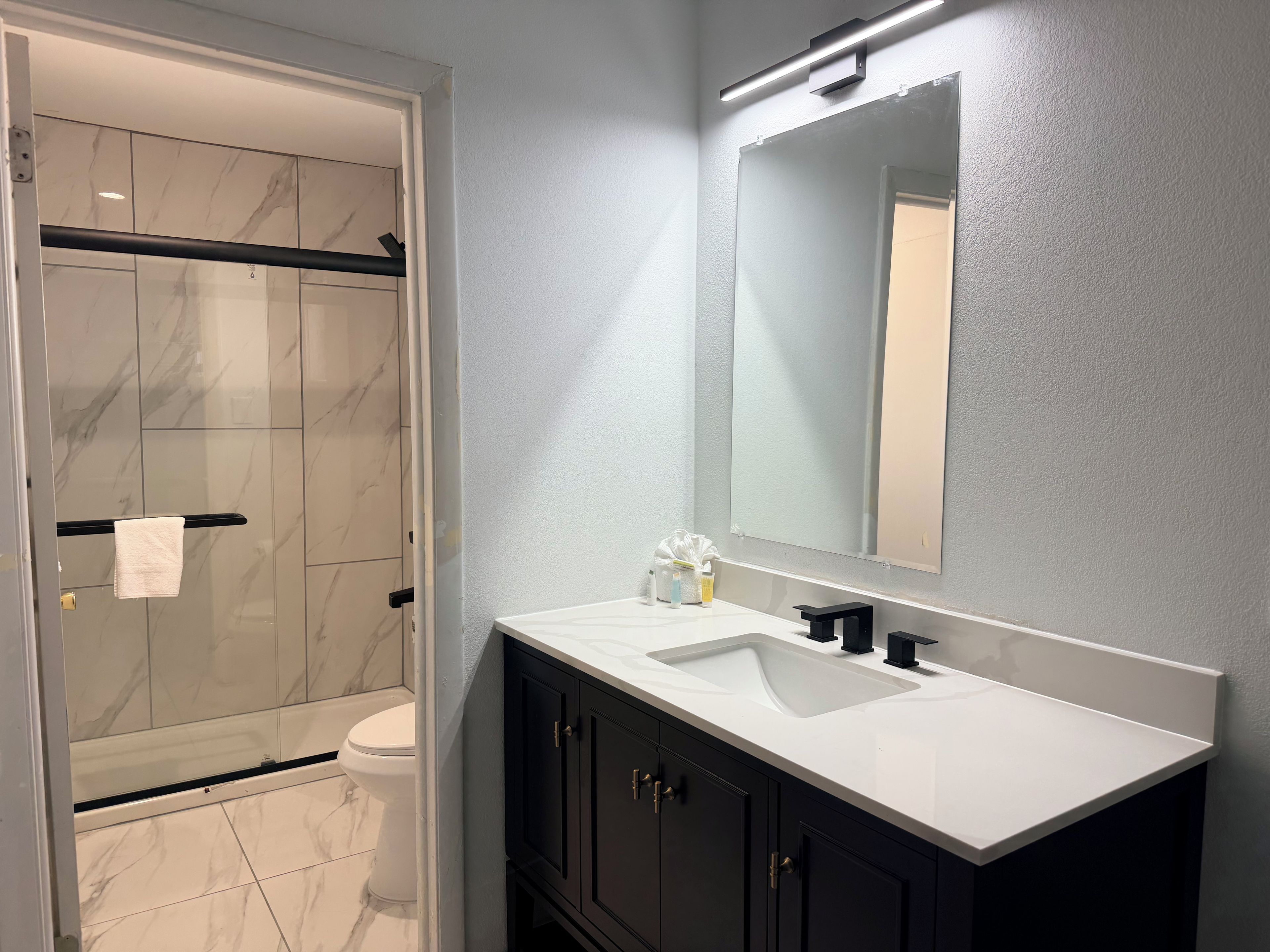 A modern bathroom with a marble shower, a white sink, dark cabinets, and a large mirror.