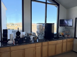 A kitchen with a large window, a coffee machine, a refrigerator, and a television mounted on the wall.