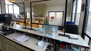 A reception desk with a computer, phone, and various papers. There are chairs and tables in the background, and a large mirror above the desk.