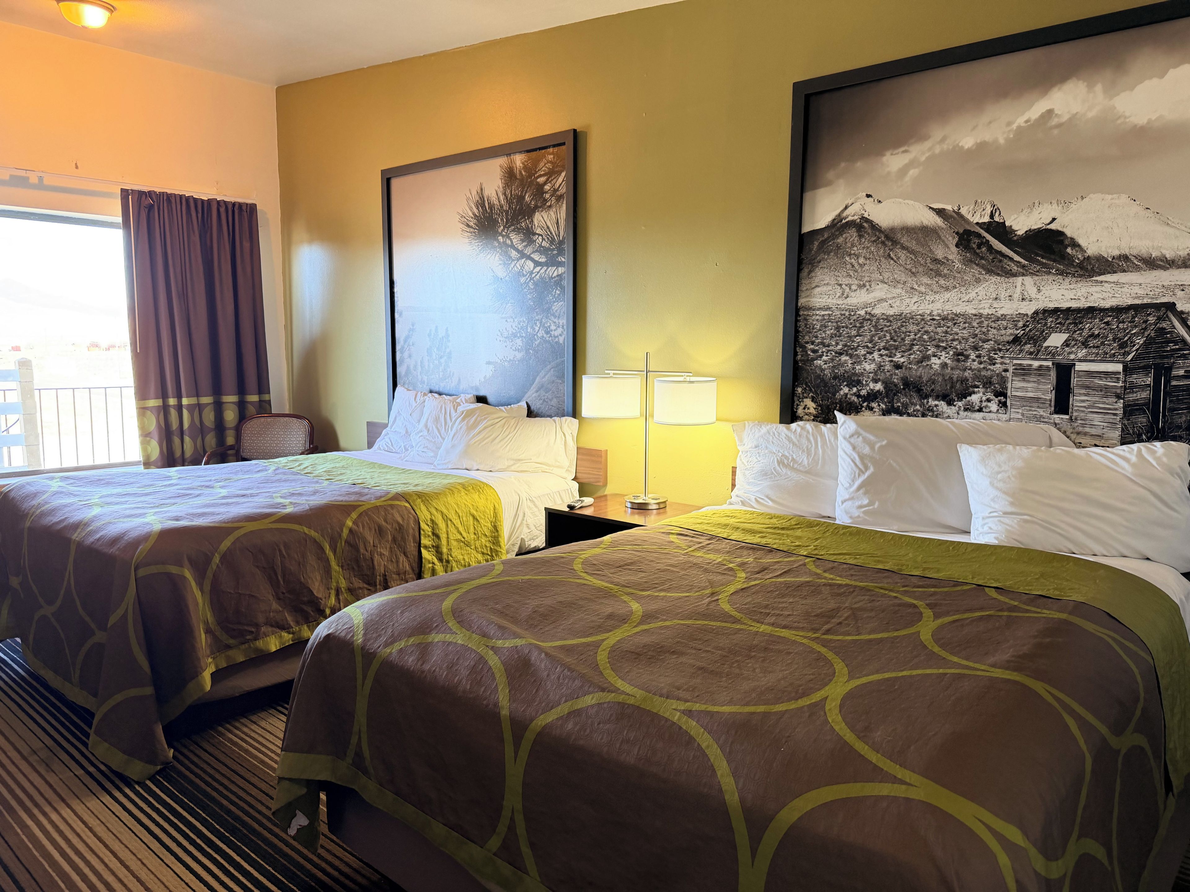 Une chambre d’hôtel avec deux lits, une grande fenêtre avec des rideaux, une chaise, une table de chevet avec une lampe et deux tableaux encadrés au mur.