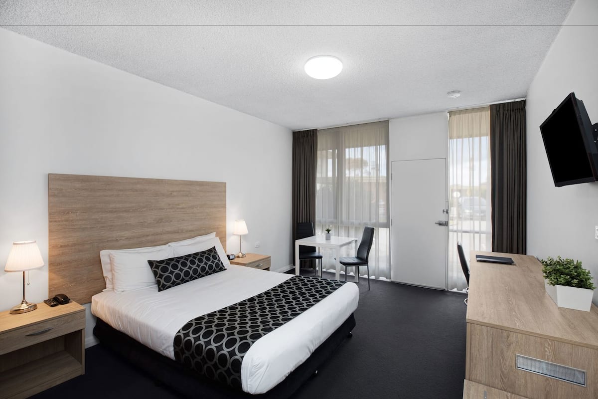 Ett hotellrum med en säng, sänglampor, ett skrivbord, en stol, en tv och ett fönster med gardiner.