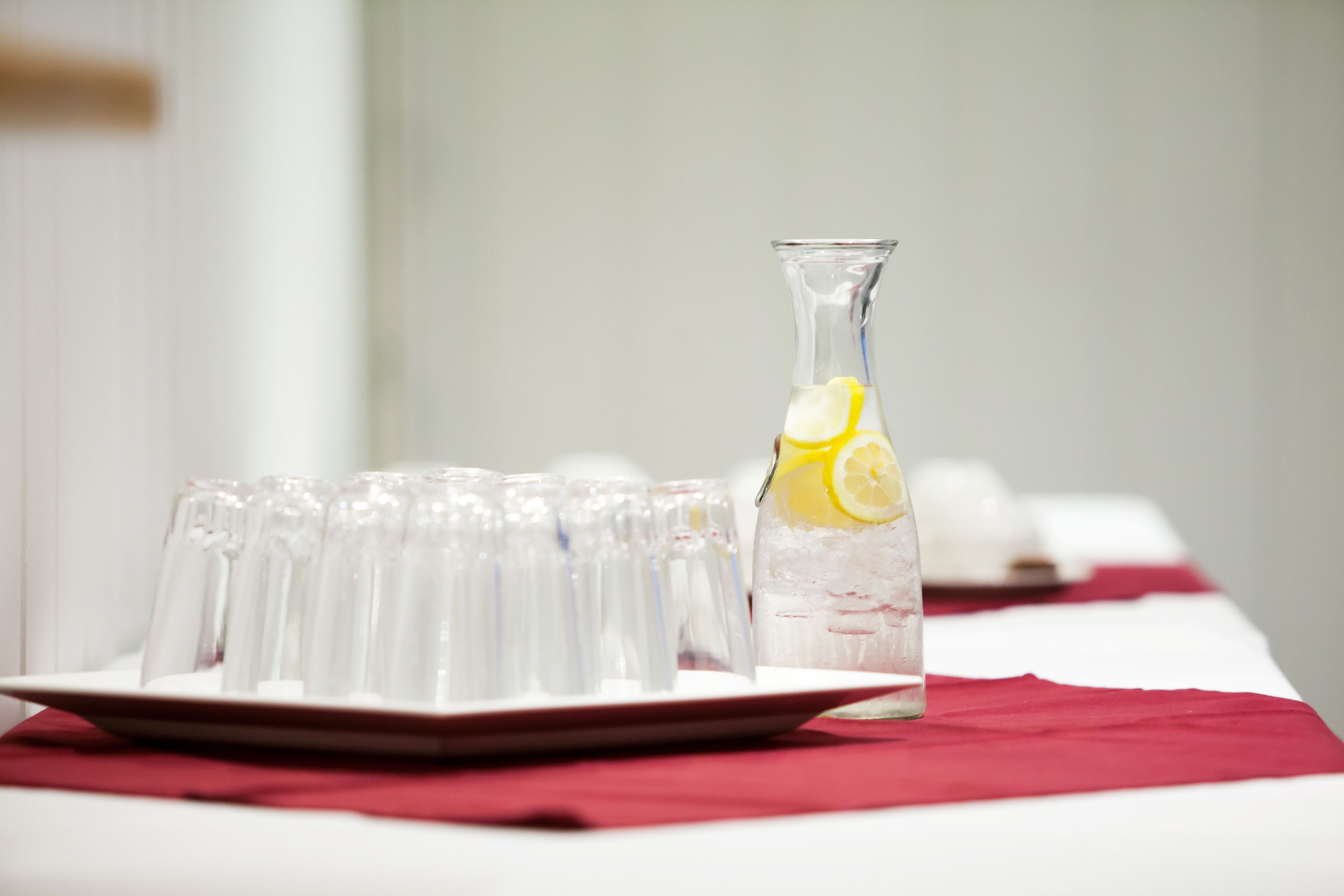 Une table sur laquelle se trouve une carafe en verre transparent remplie d’une boisson contenant des tranches de citron, entourée de verres vides posés sur une nappe rouge.