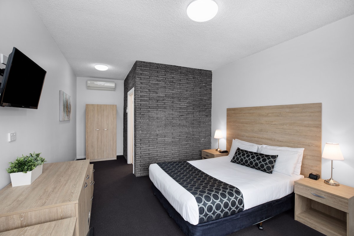 Ett modernt hotellrum med en stor säng, sängbord, en platt-TV som är monterad på väggen och ett litet skrivbord med lådor.