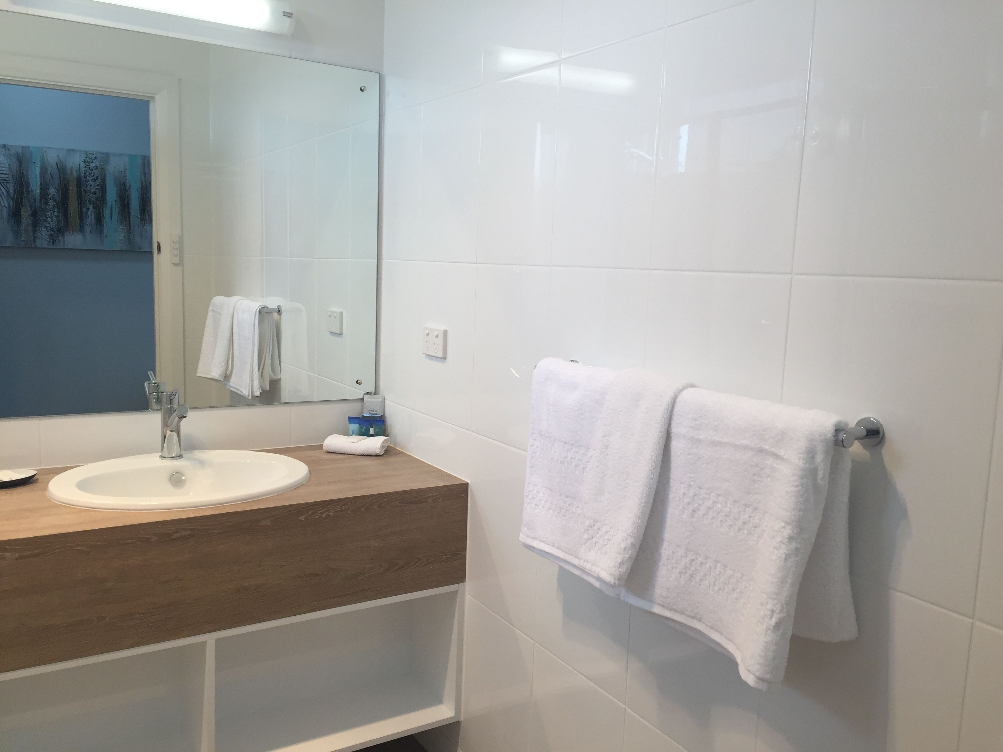 Un baño moderno con tocador de madera, lavamanos blanco y un espejo grande. Hay dos toallas blancas colgadas en un perchero.