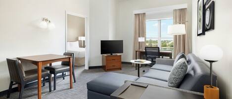A hotel room with a dining area, a sofa, a TV, and a window with curtains.