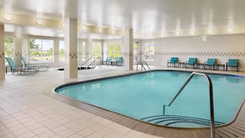 Una piscina interior con suelo de baldosas, sillas de descanso y grandes ventanales.