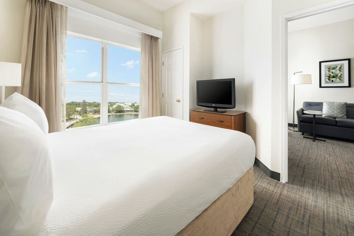 A hotel room with a large bed, a television on a wooden stand, a desk with a lamp, and a view of a body of water and greenery.