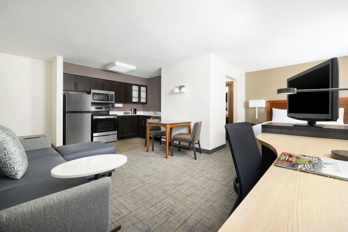 A modern hotel room with a kitchenette, a dining area, a sofa, and a flat-screen TV.