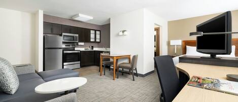 A modern hotel room with a kitchenette, a dining area, a sofa, and a flat-screen TV.