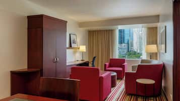 Habitación de hotel con un armario grande, una mesa de comedor, sillas tapizadas rojas, una pequeña mesa redonda y vistas de la ciudad a través de ventanas amplias.