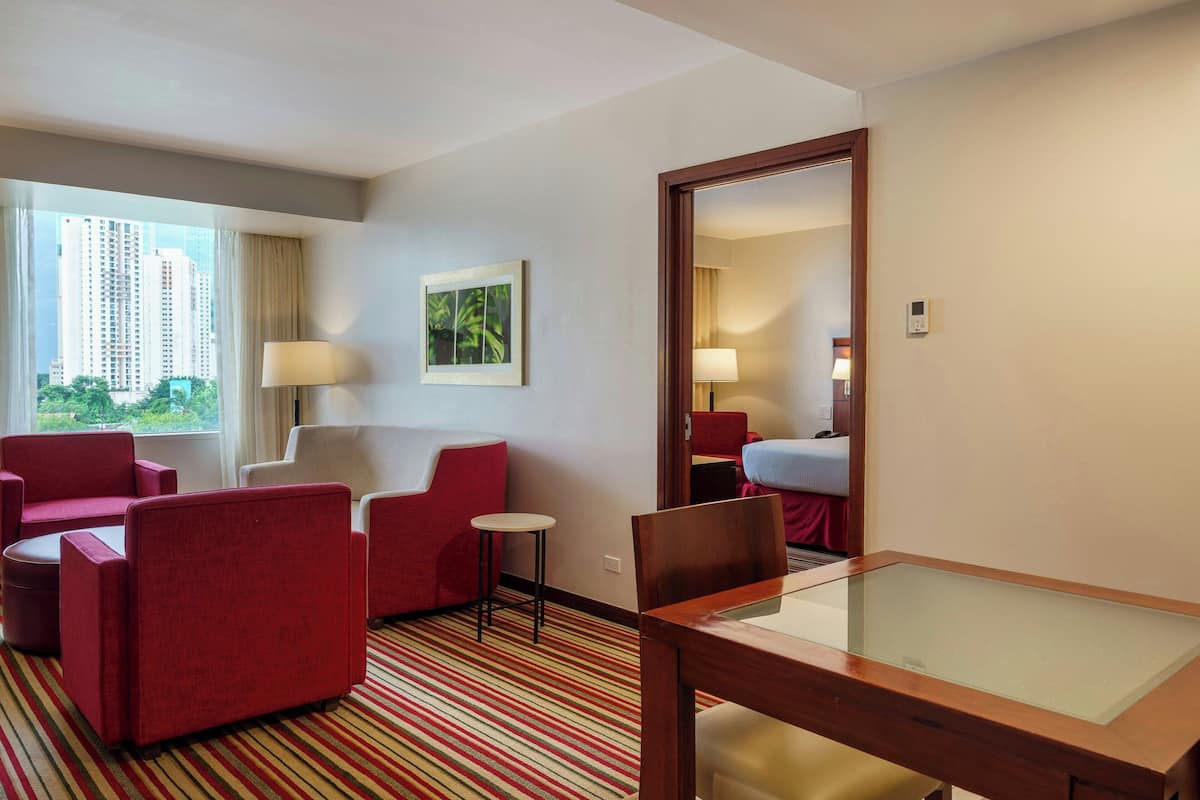 A hotel room with a large window, red upholstered chairs, a small round table, a glass-top table, and a mirror on the wall.