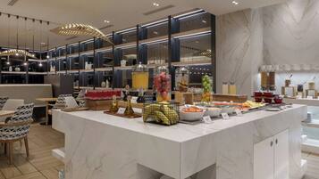 Un área de desayuno de hotel moderna con mostradores de mármol, diversas frutas y bebidas, y mobiliario contemporáneo.