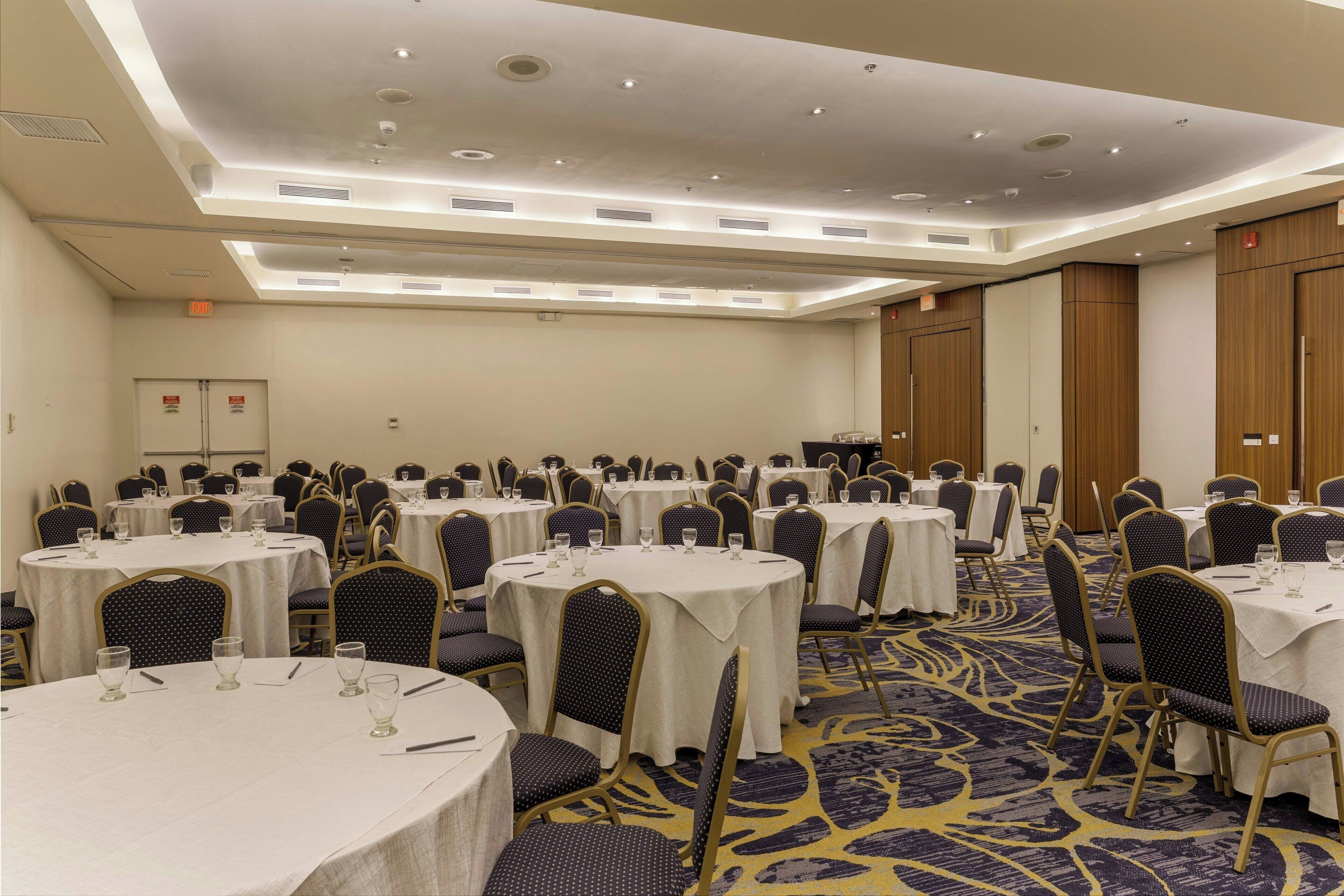 Un salón de conferencias de hotel con varias mesas redondas, sillas y piso alfombrado.