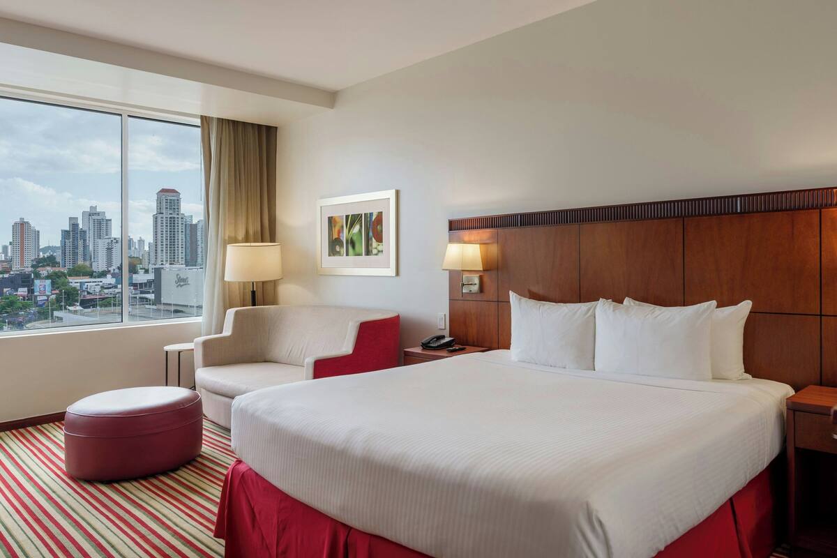 A hotel room with a large bed, a chair, a small ottoman, a nightstand, a lamp, and a city view through the window.