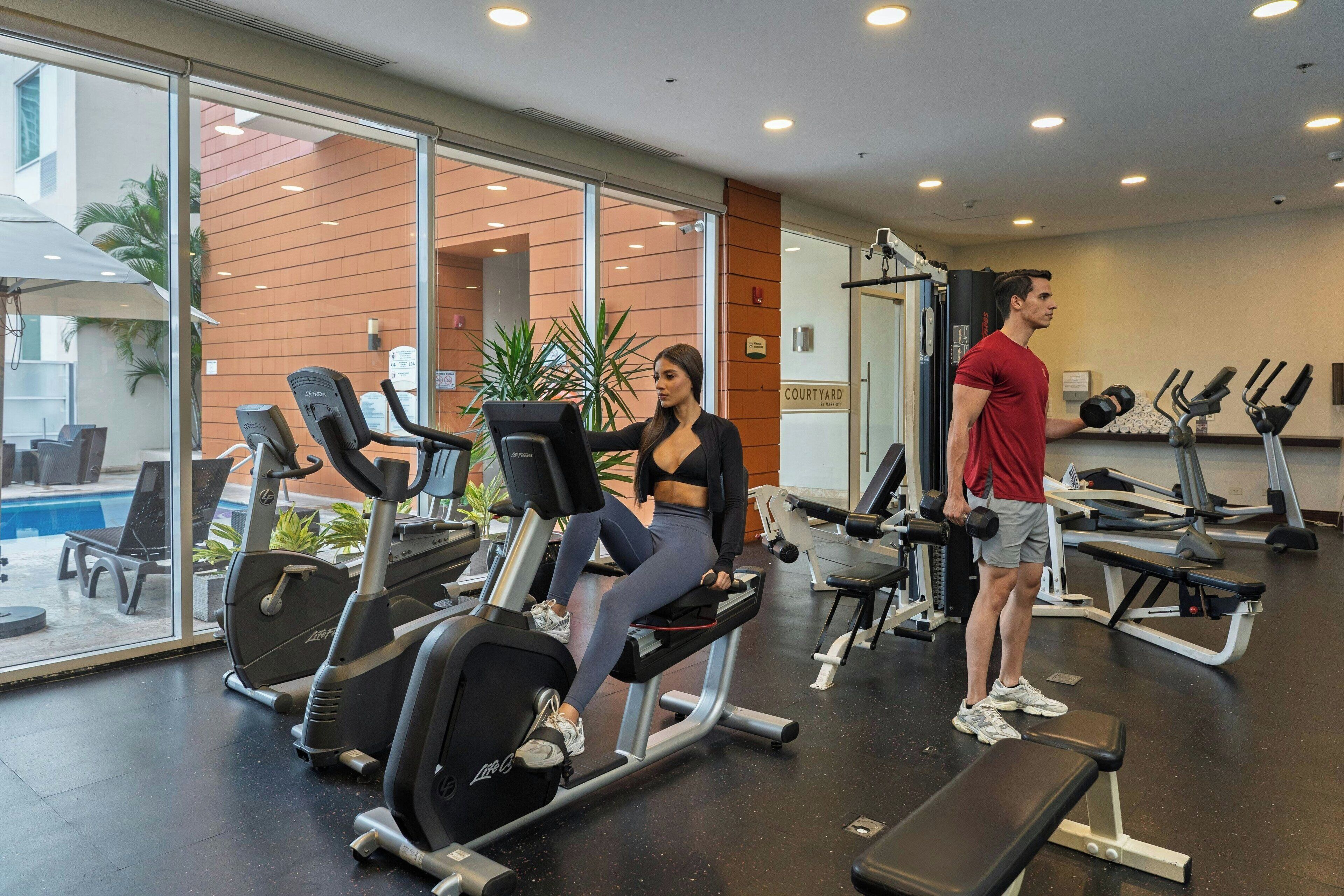 A gym with cardio equipment, benches, and a pool visible through the glass wall.