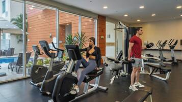 A gym with cardio equipment, benches, and a pool visible through the glass wall.