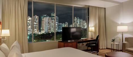 Habitación de hotel con una cama grande, un escritorio con una computadora, un televisor y vistas a un paisaje urbano nocturno.