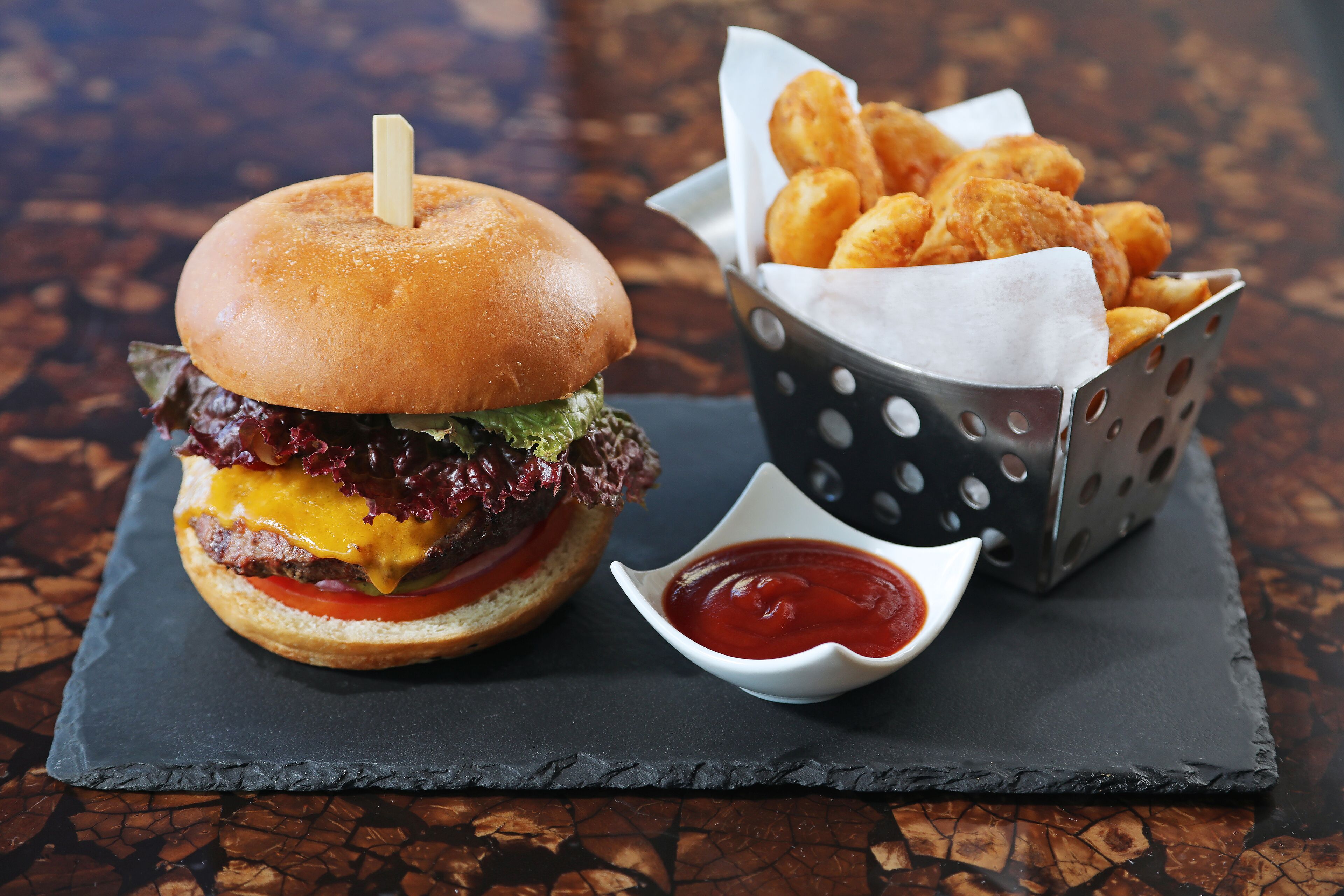 Una hamburguesa con un palillo, una cesta de aros de cebolla y un cuenco pequeño de kétchup sobre una tabla de pizarra.