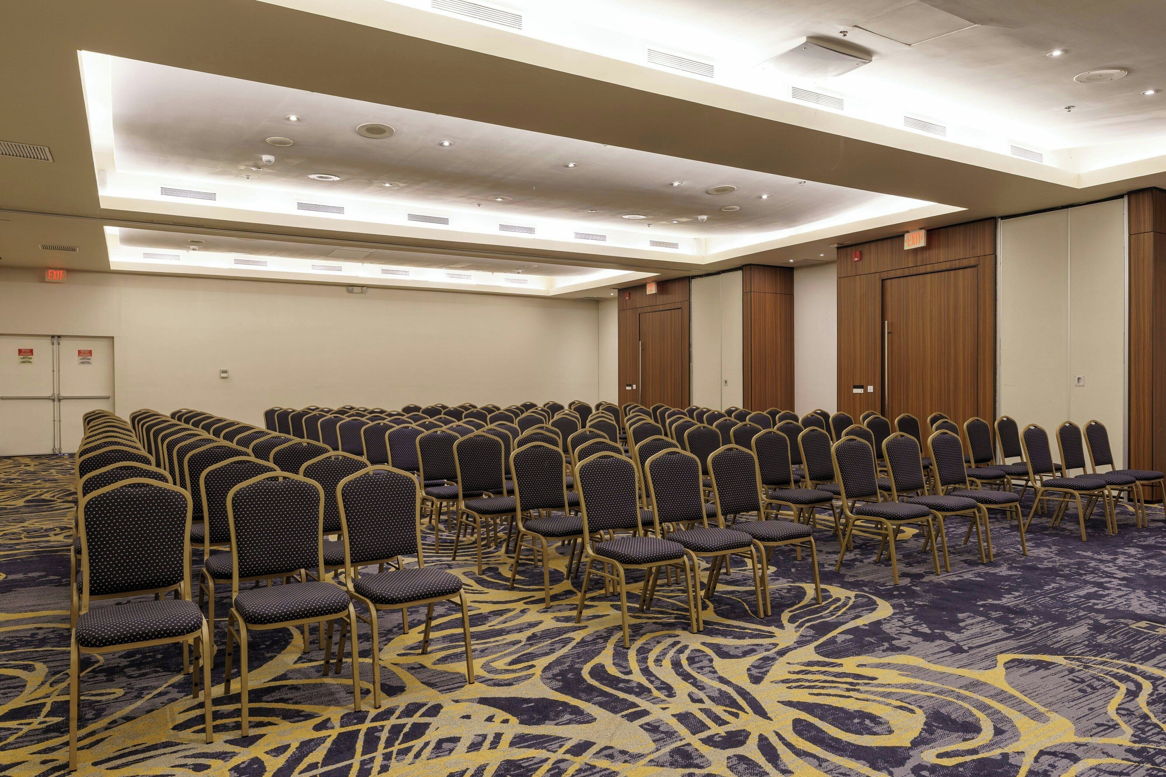 Un salón de conferencias con filas de sillas, piso alfombrado e iluminación empotrada en el techo.