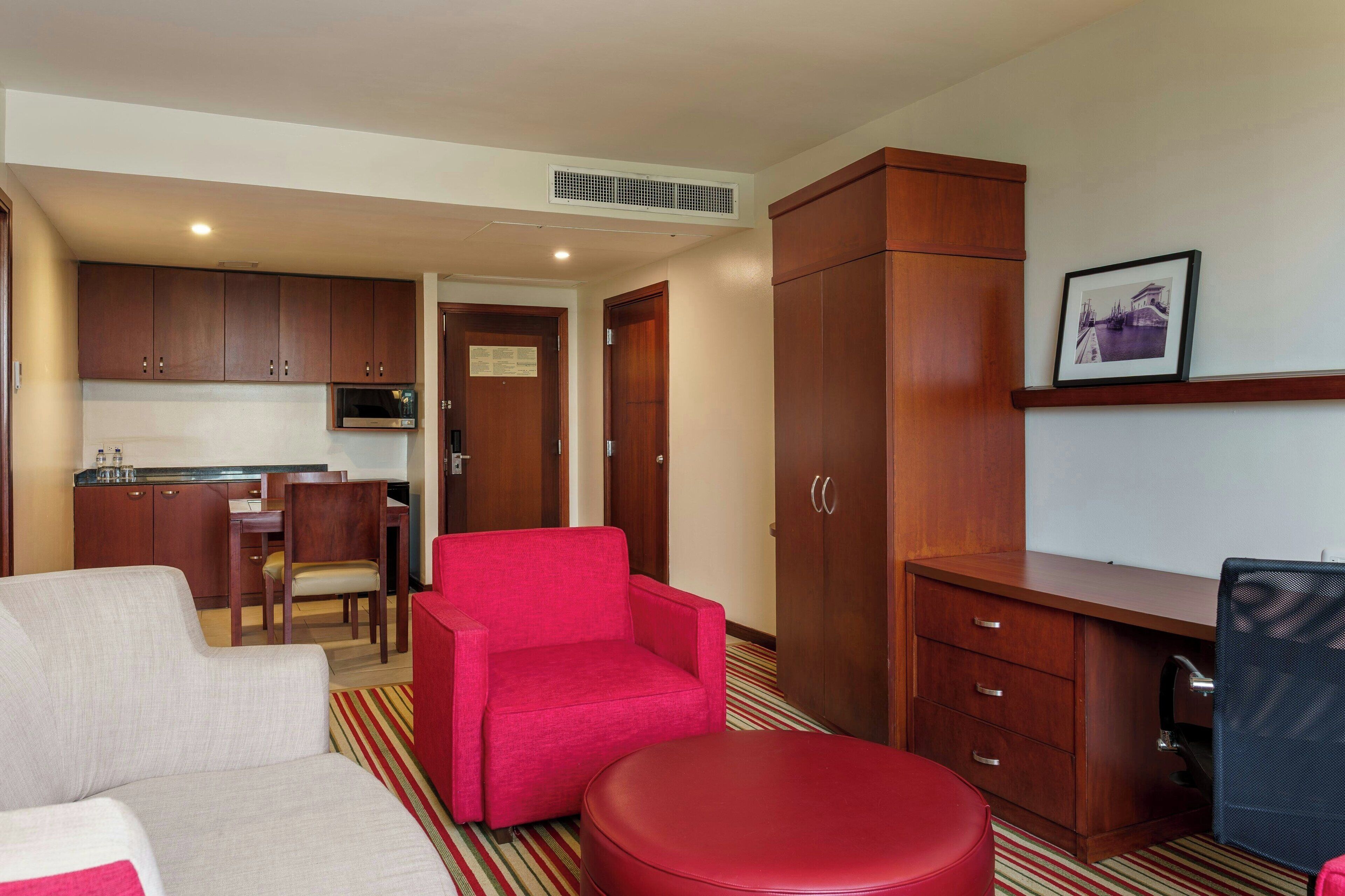 Une chambre d’hôtel compacte comprenant un coin cuisine, un fauteuil rouge, un canapé et un bureau avec une chaise.