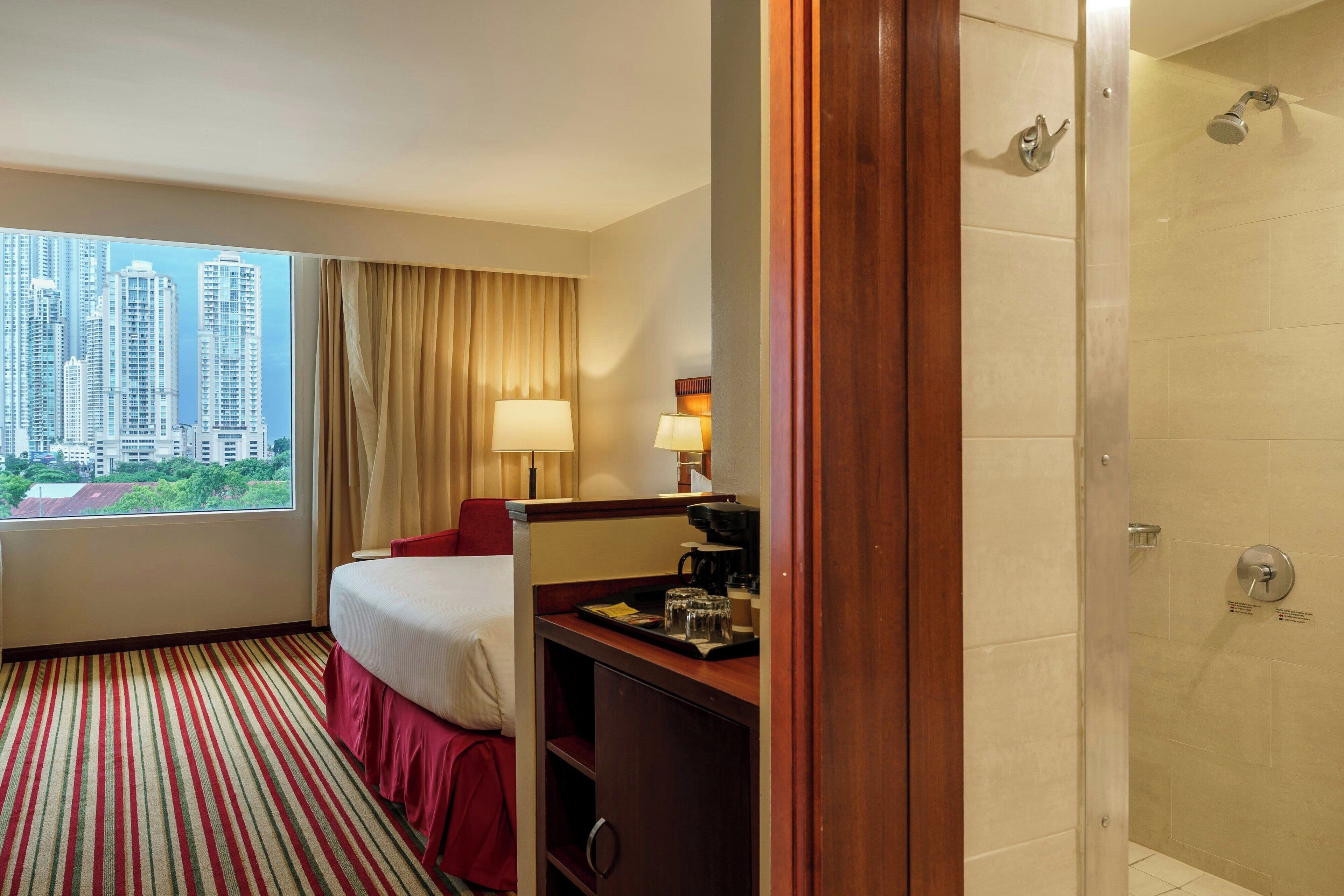 Ein Hotelzimmer mit Bett, Schreibtisch mit Lampe, einer Kaffeemaschine und Blick auf hohe Gebäude.