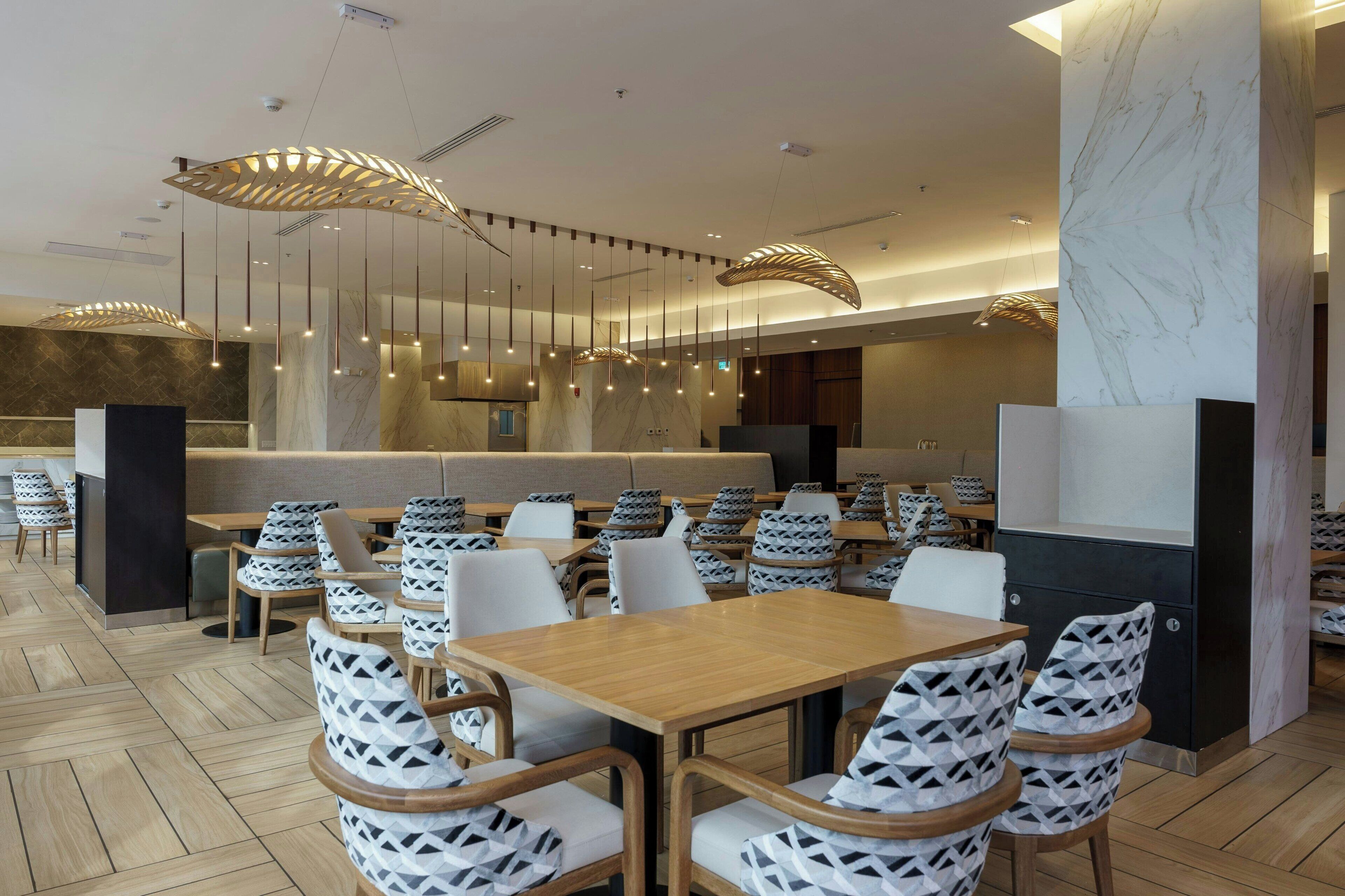 Um restaurante moderno com mesas de madeira e cadeiras estampadas, colunas de mármore e luminárias contemporâneas.