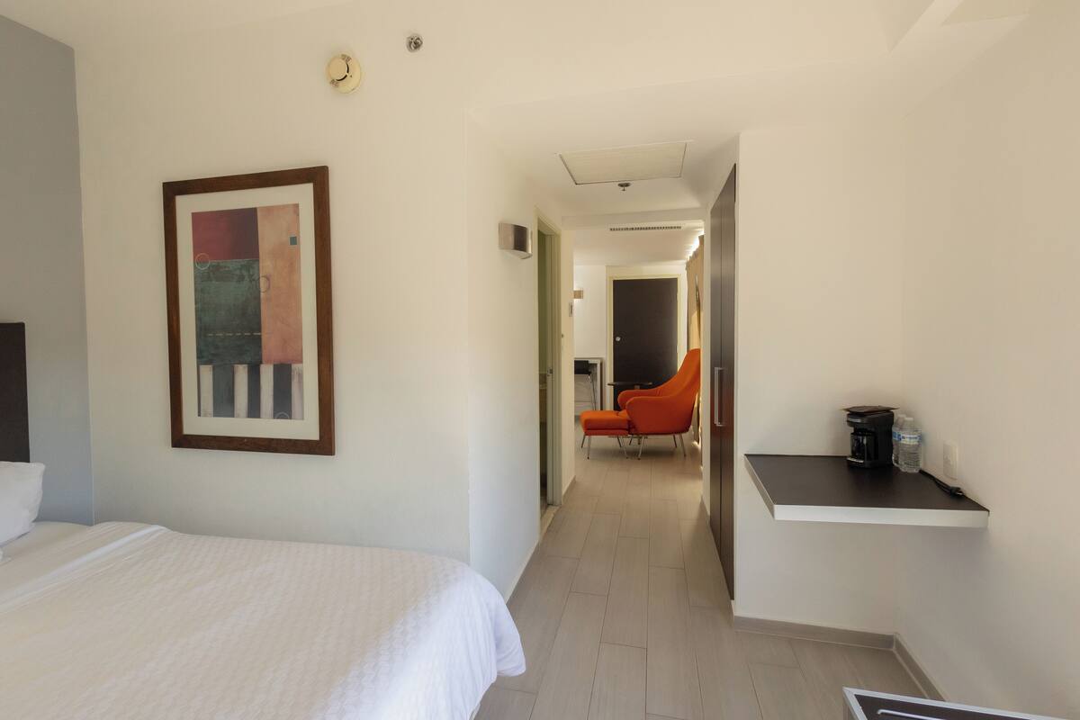 A hotel room with a bed, a framed abstract painting, a chair, and a small table with a coffee maker.