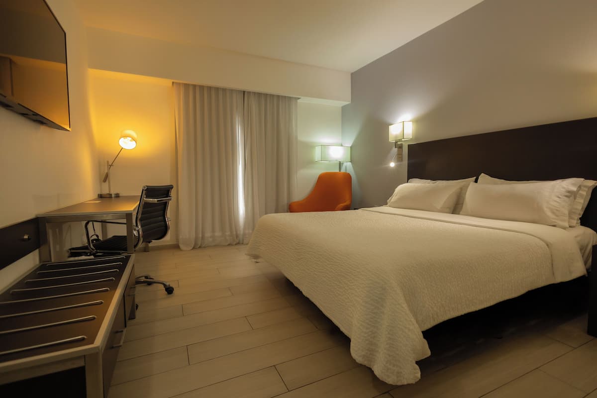 A hotel room with a bed, desk, chair, and a lamp.