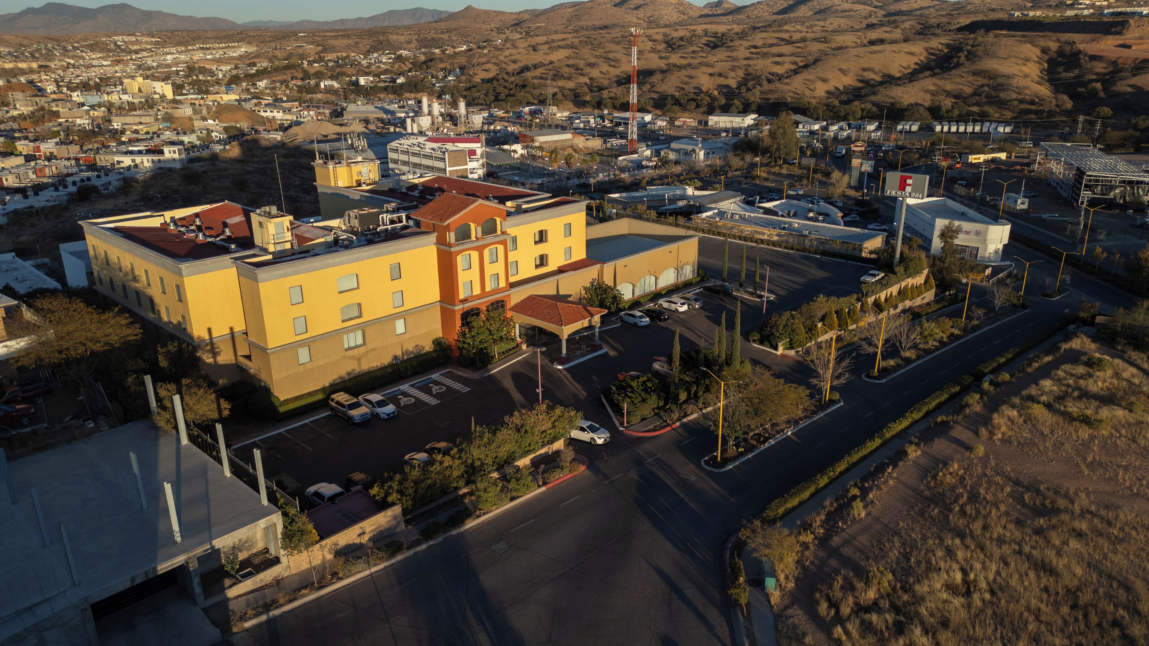 An aerial view of a hotel with a parking lot, a small plaza, and a mountainous backdrop.