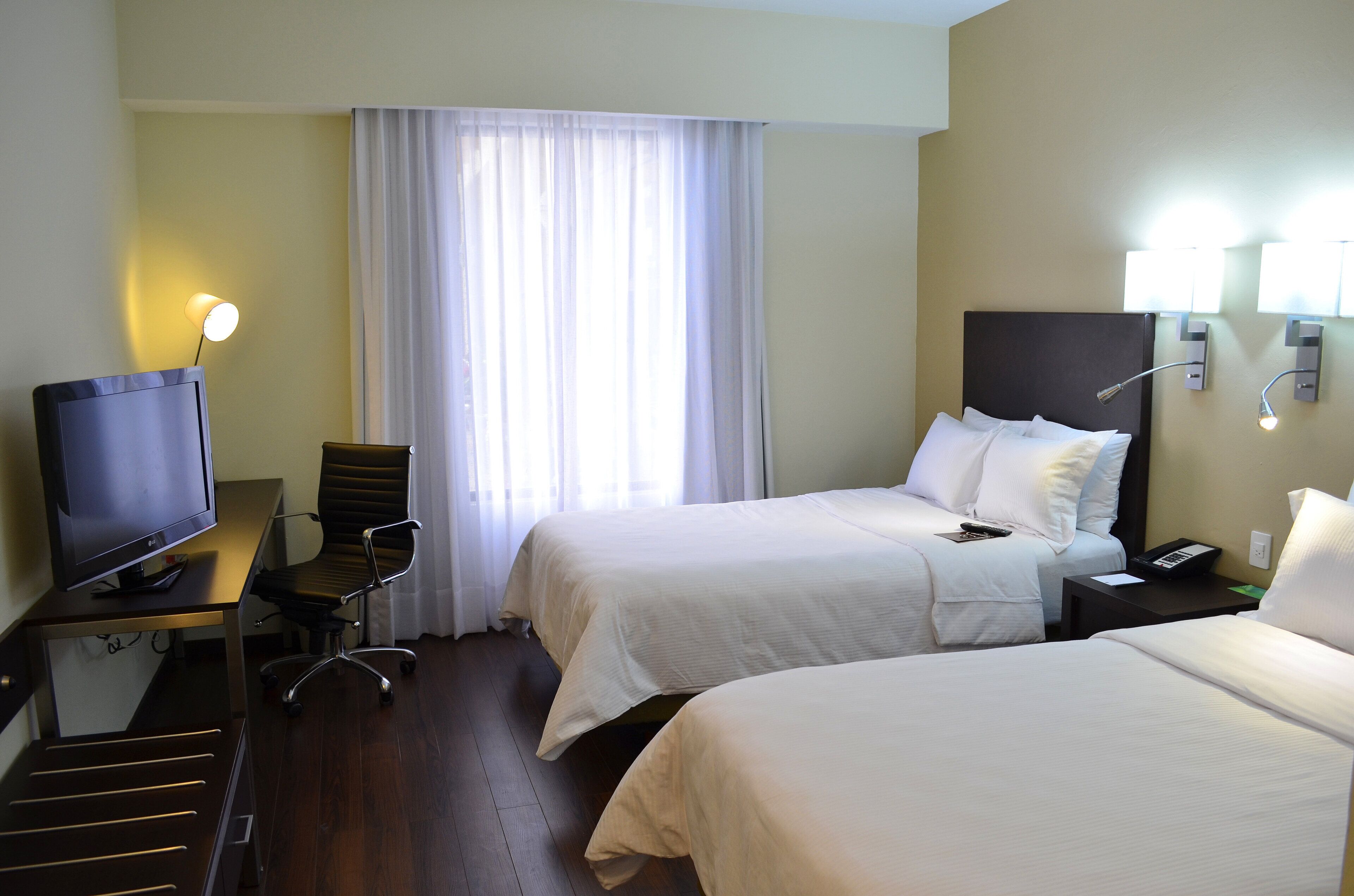 Camera d'albergo con due letti, una scrivania con televisore, una sedia e una finestra con tende.