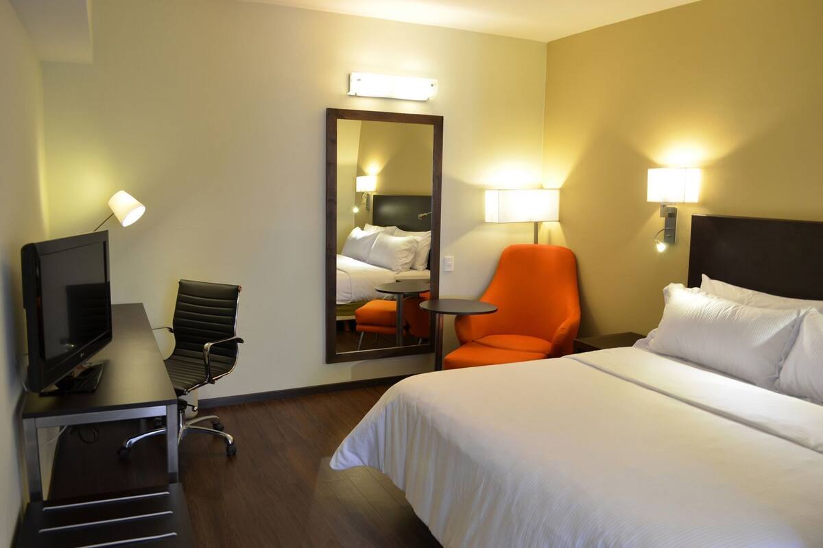 A hotel room with a bed, a desk with a TV, an orange chair, a small table, and a mirror.