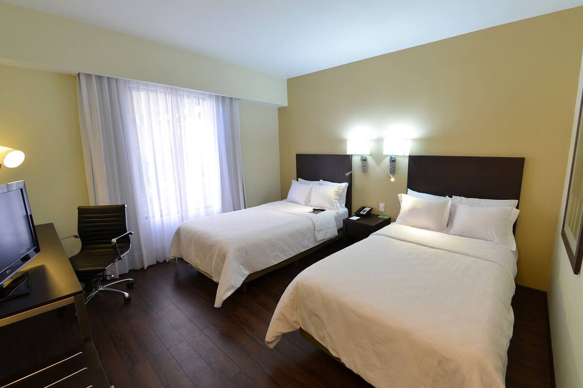 A hotel room with two beds, a desk with a computer, a chair, and a window with curtains.