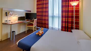 A hotel room with a bed, a desk with a computer, a chair, a television, and a teddy bear on the bed.