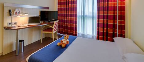 Habitación de hotel con una cama, un escritorio con computadora, una silla, un televisor y un oso de peluche sobre la cama.