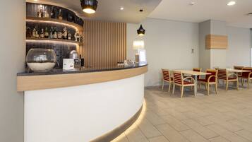 A modern bar area with a curved counter, various bottles, a coffee machine, and a bowl of pebbles. There are wooden chairs and tables arranged for seating.