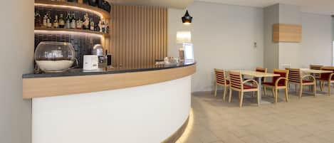 Zona de bar moderna con una barra curva, varias botellas, una máquina de café y un cuenco de guijarros. Hay sillas y mesas de madera dispuestas para sentarse.