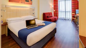 Quarto de hotel com uma cama grande, uma escrivaninha com um laptop, uma poltrona vermelha e vista da cidade através de janelas amplas.