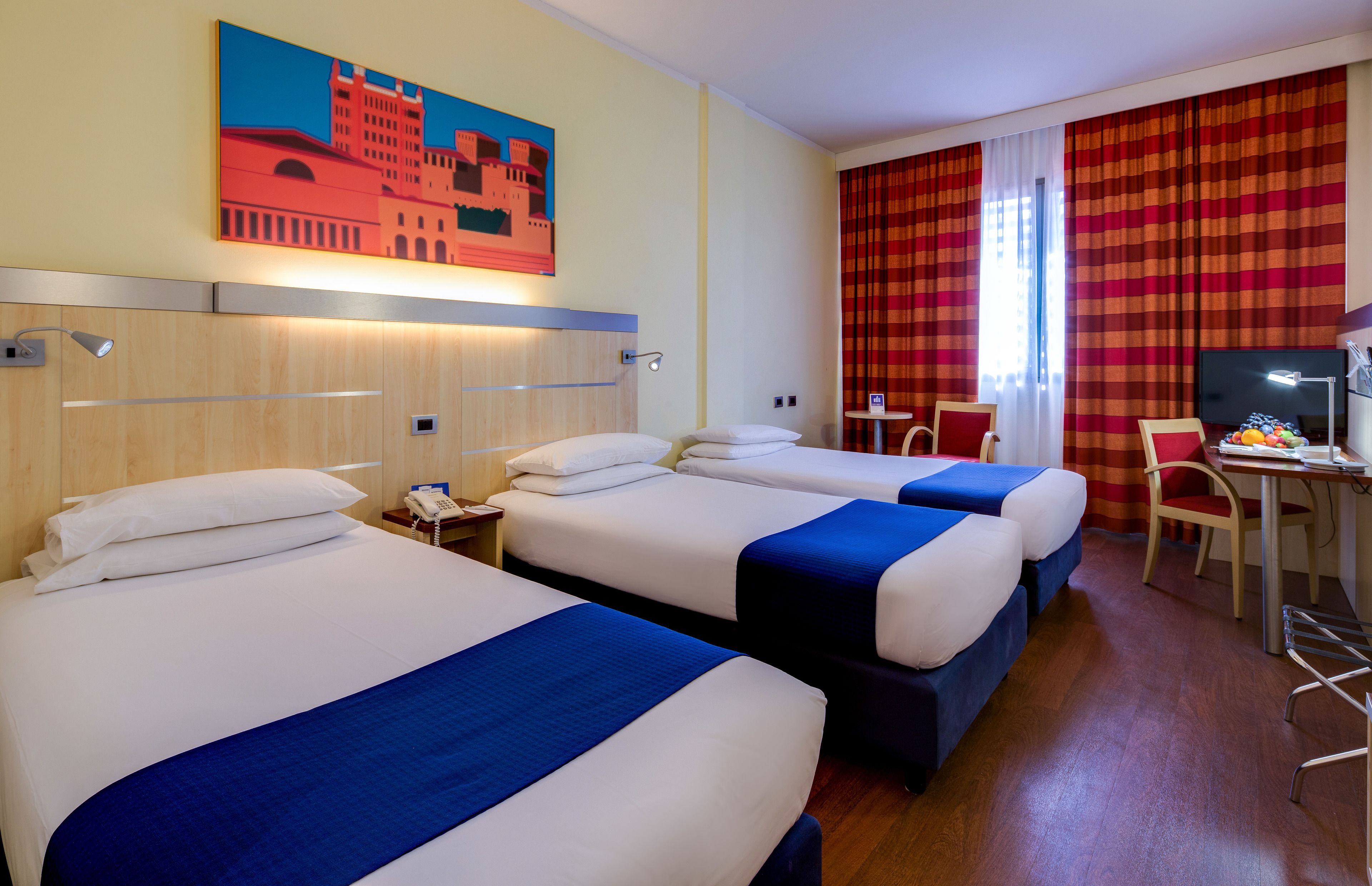 A hotel room with three beds, a desk with a chair, a television, and a painting on the wall.