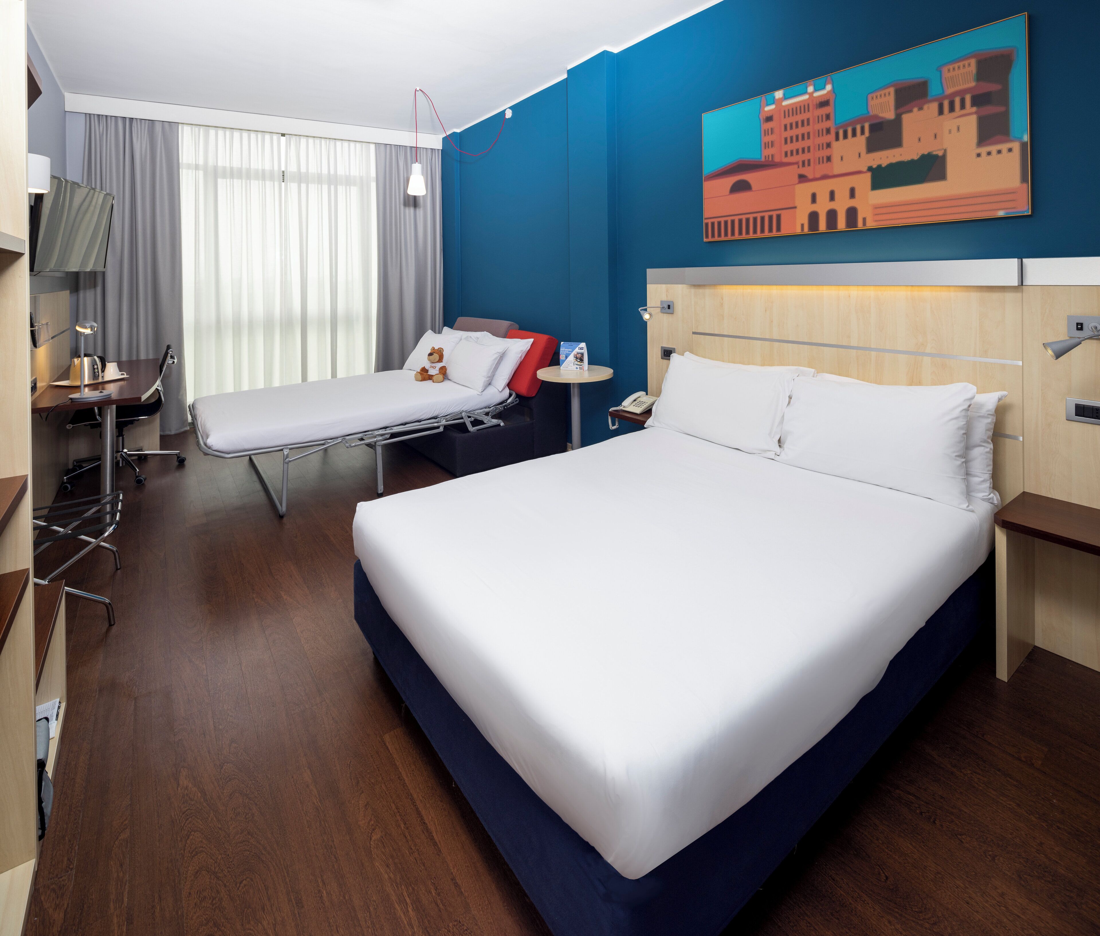 Habitación de hotel con una cama grande, una cama más pequeña, un escritorio con silla, una mesita de noche, una lámpara y un cuadro en la pared.
